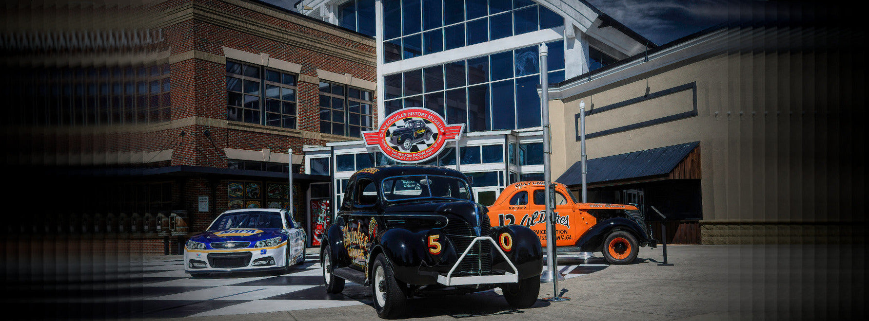 A black vintage custom car, parked in front of the Georgia Racing Hall of Fame. An orange vintage car and Chase Elliott's NAPA car are partially visible in the background.